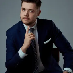 Portret mężczyzny w granatowym garniturze, białej koszuli i szarym krawacie, siedzącego na krześle i zamyślonego, z palcem wskazującym uniesionym do góry.