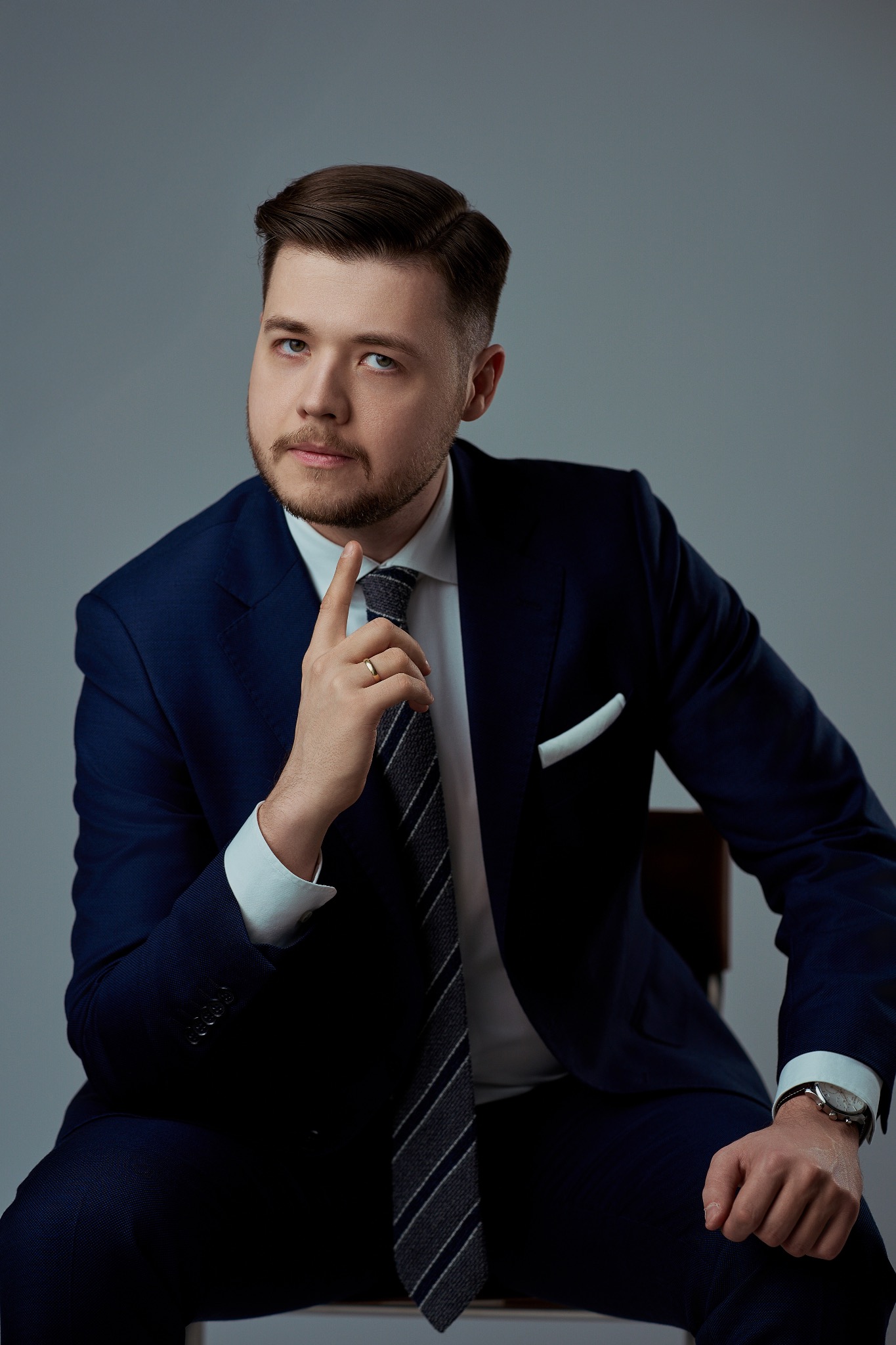 Portret mężczyzny w granatowym garniturze, białej koszuli i szarym krawacie, siedzącego na krześle i zamyślonego, z palcem wskazującym uniesionym do góry.