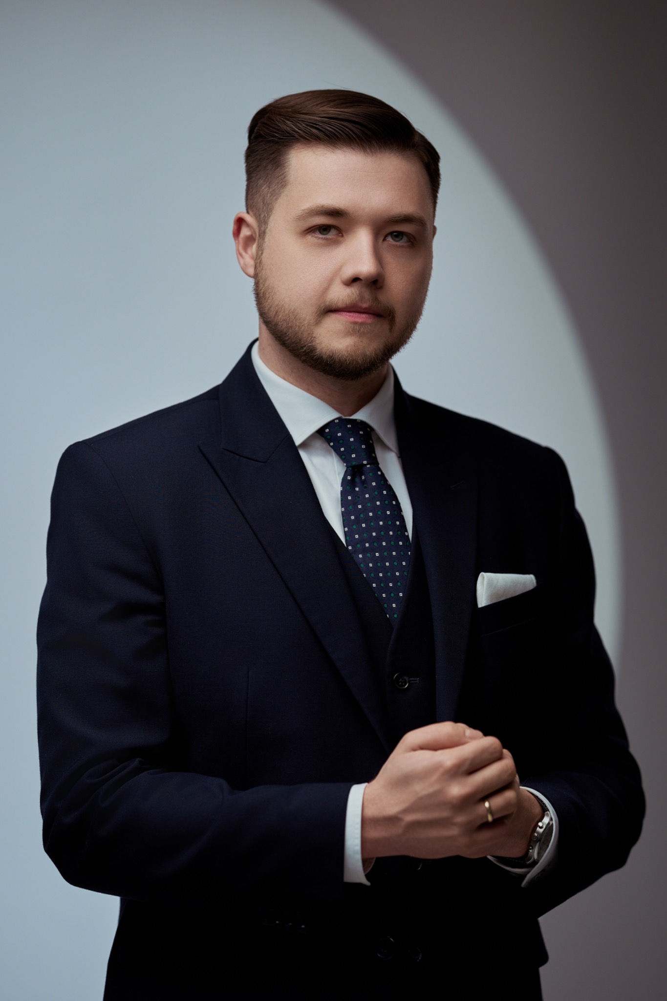 Portret mężczyzny w granatowym garniturze, białej koszuli i wzorzystym krawacie, z delikatnym uśmiechem i splecionymi dłońmi, na jasnoszarym tle.