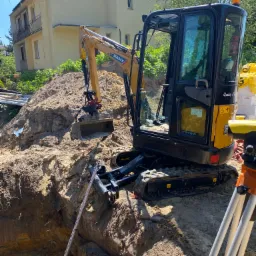 Żółto-czarna mini koparka SANY na gąsienicach stoi obok wykopu w ziemi, w tle budynek mieszkalny i drzewa, widoczna łata niwelacyjna i niwelator optyczny.