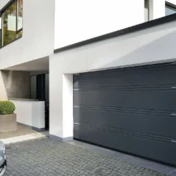 Nowoczesna posesja z szarą bramą garażową segmentową, srebrnym autem i kostką brukową w szarym odcieniu.