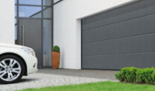 Nowoczesna, segmentowa brama w kolorze antracytowym, z poziomymi przetłoczeniami, zamontowana w jasnym budynku z szarą stolarką okienną i drzwiami wejściowymi. Fragment białego samochodu widoczny...