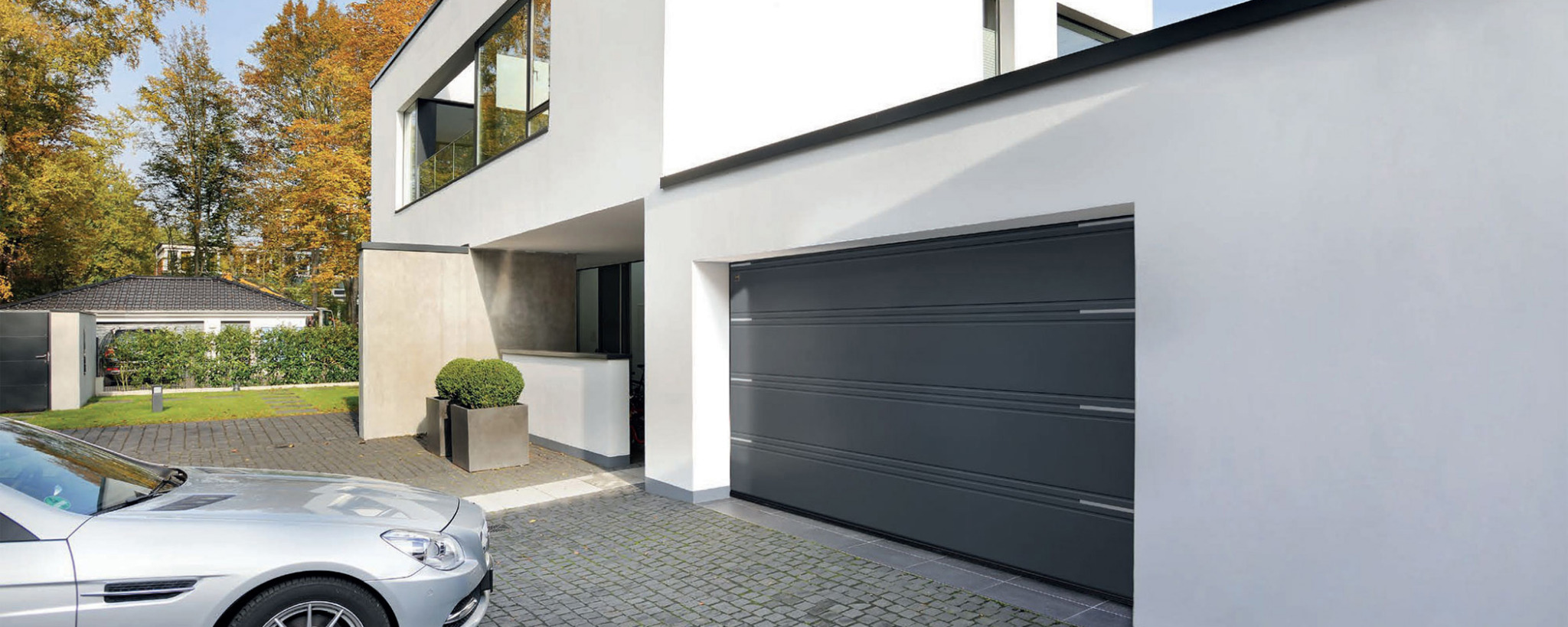 Nowoczesna posesja z szarą bramą garażową segmentową, srebrnym autem i kostką brukową w szarym odcieniu.