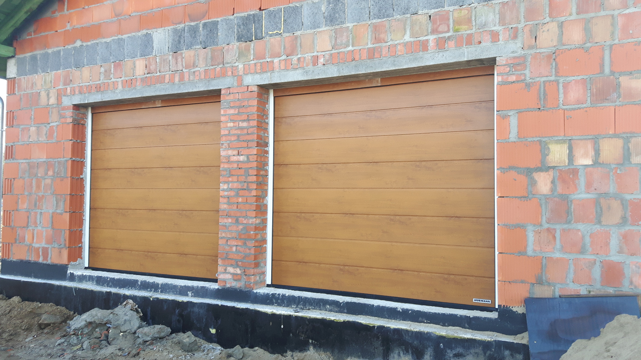 Dwusegmentowa brama garażowa w kolorze drewna, zamontowana w surowym murze z cegły, widoczne logo producenta w lewym dolnym rogu.