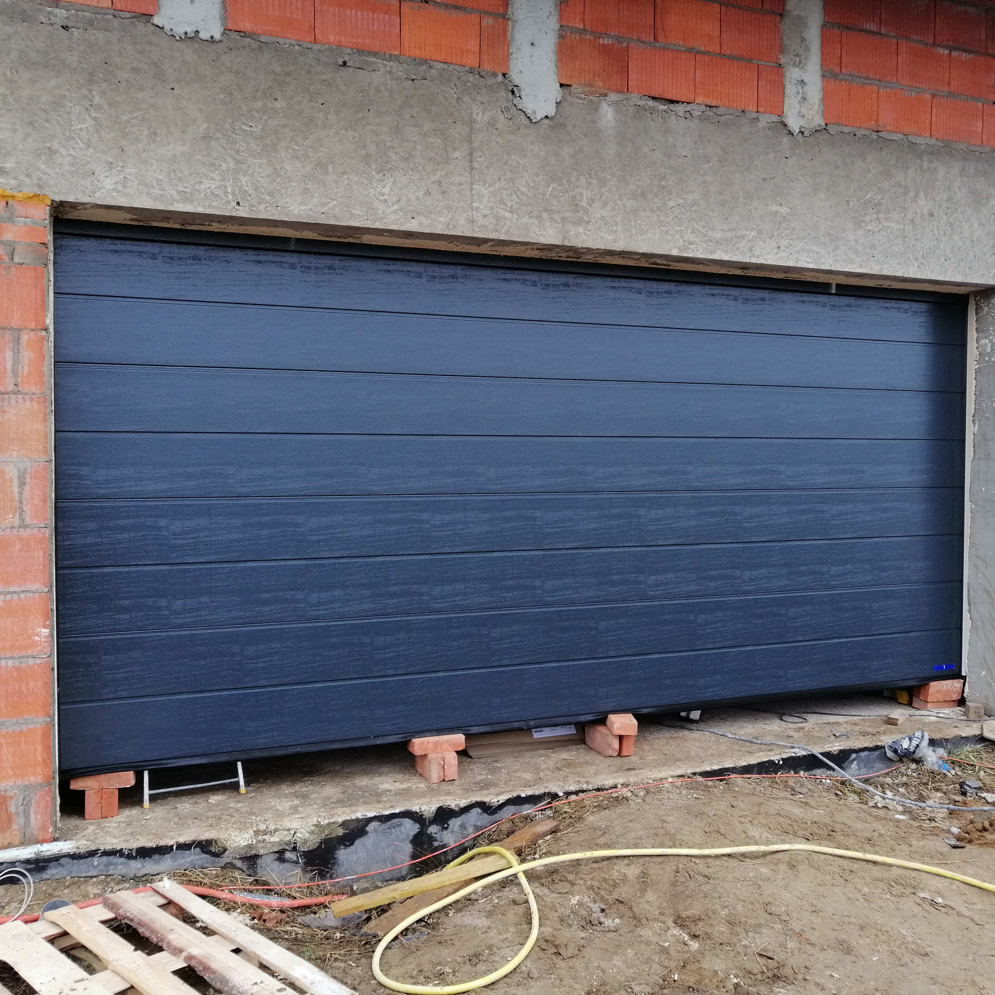 Nowa, segmentowa brama garażowa w kolorze antracytowym, tymczasowo podparta cegłami podczas montażu w surowym stanie budynku z widoczną instalacją elektryczną w ziemi.