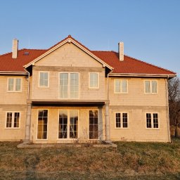 WILK.ins - Surowy stan dwupiętrowego domu z czerwonym dachem i białymi oknami, w trakcie budowy, z widocznymi pustakami i nieotynkowanymi ścianami, na tle błękitnego nieba o zachodzie słońca.