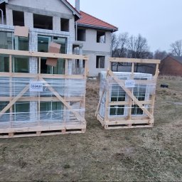 WILK.ins - Dwa drewniane palety z zapakowanymi oknami PCV, zabezpieczone folią, stoją na trawie przed budynkiem w trakcie budowy.