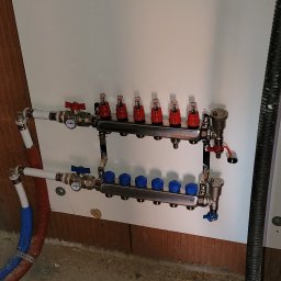 WILK.ins - Rozdzielacz do ogrzewania podłogowego z czerwonymi i niebieskimi zaworami, zamontowany na ścianie w surowym wnętrzu.