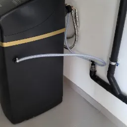 Czarny zmiękczacz wody z żółtymi akcentami, podłączony do instalacji wodnej za pomocą elastycznych węży i rur PCV. Widoczny filtr na rurze odpływowej.