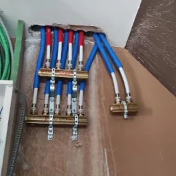 Rozdzielacz ogrzewania podłogowego z rurami PEX w kolorach: niebieskim, białym i czerwonym, zamontowany na ścianie w trakcie instalacji. Widoczne elementy mocujące.