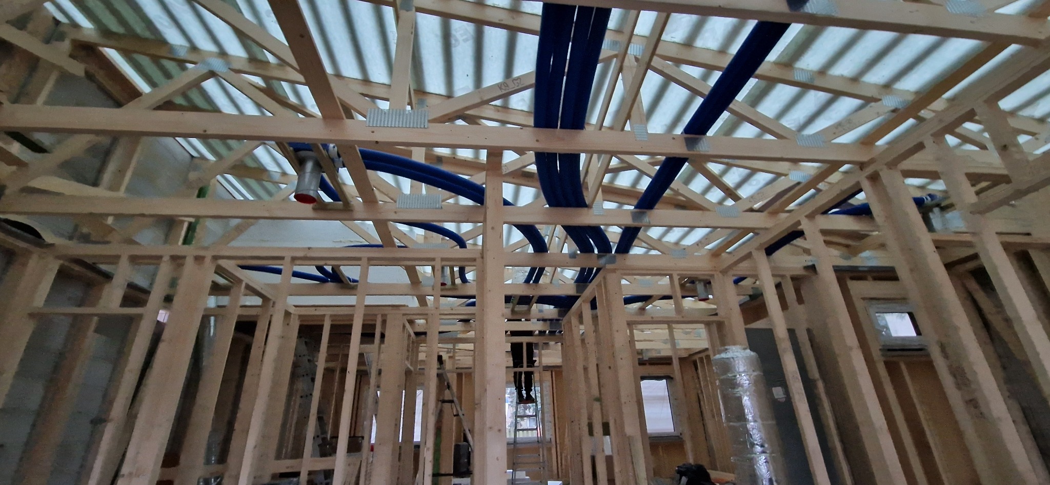 Wnętrze domu w trakcie budowy z widoczną drewnianą konstrukcją szkieletową i rozprowadzonymi niebieskimi rurami wentylacyjnymi pod sufitem, mocowanymi za pomocą metalowych uchwytów.