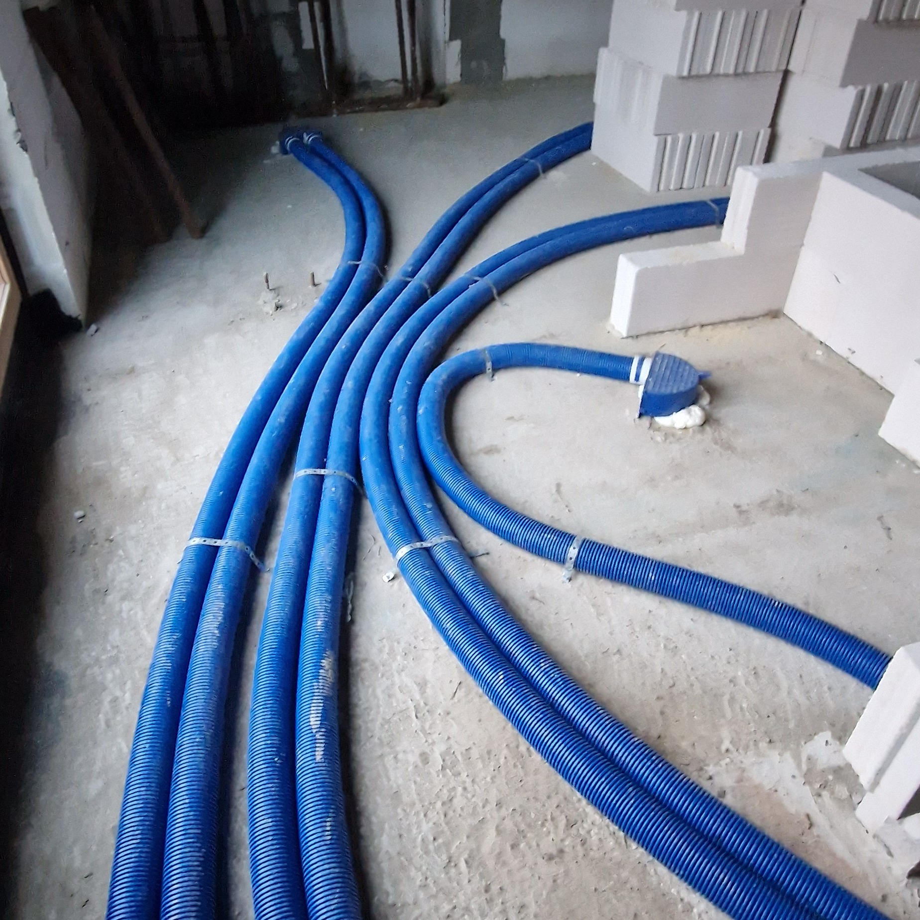 Niebieskie, karbowane rury wentylacyjne ułożone na surowym, betonowym podłożu w trakcie montażu, z widocznymi elementami mocującymi i fragmentami białych bloczków konstrukcyjnych w tle.