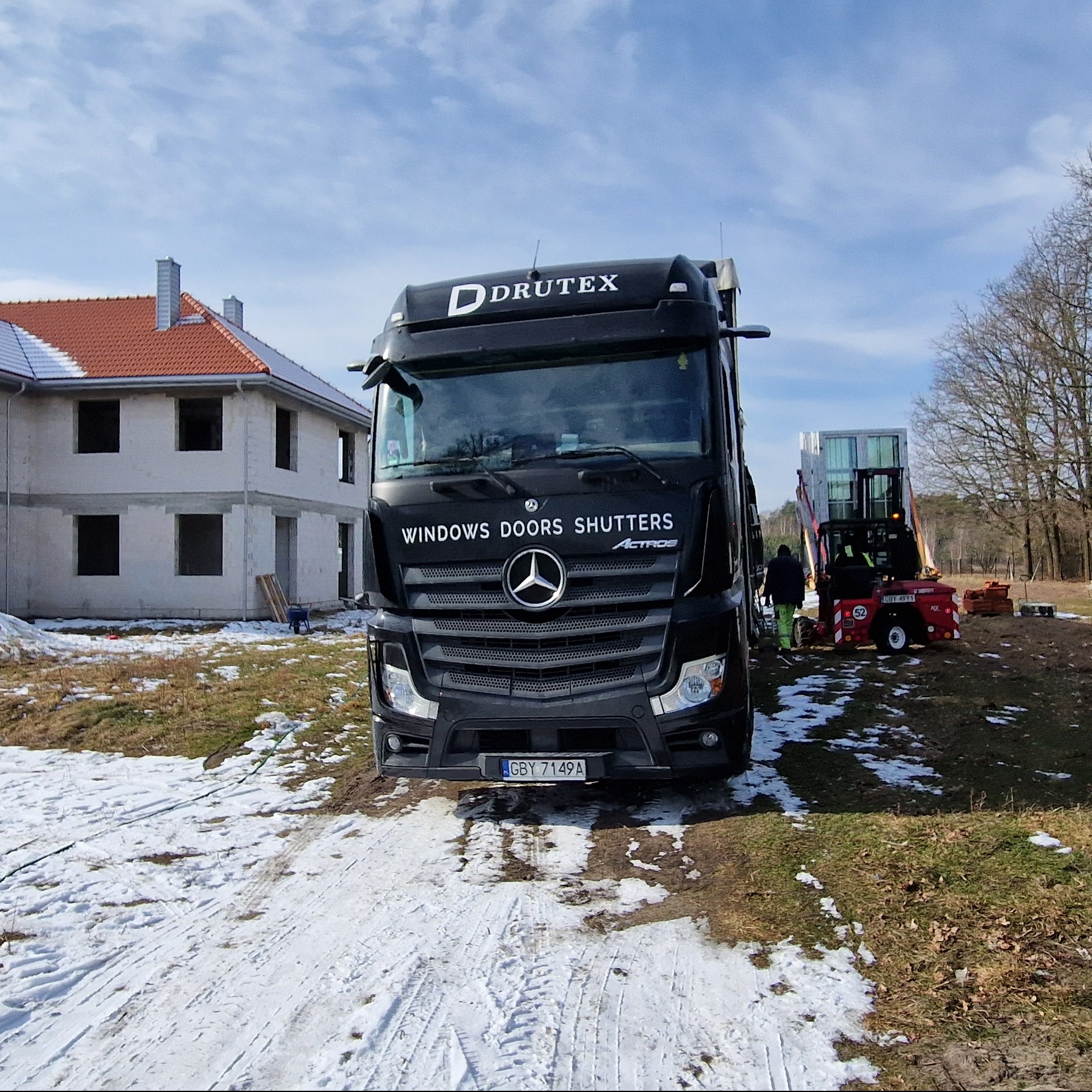 Czarna ciężarówka firmy Drutex z napisem WINDOWS DOORS SHUTTERS na tle budowanego domu w Łodzi, w tle widać wózek widłowy z oknami.