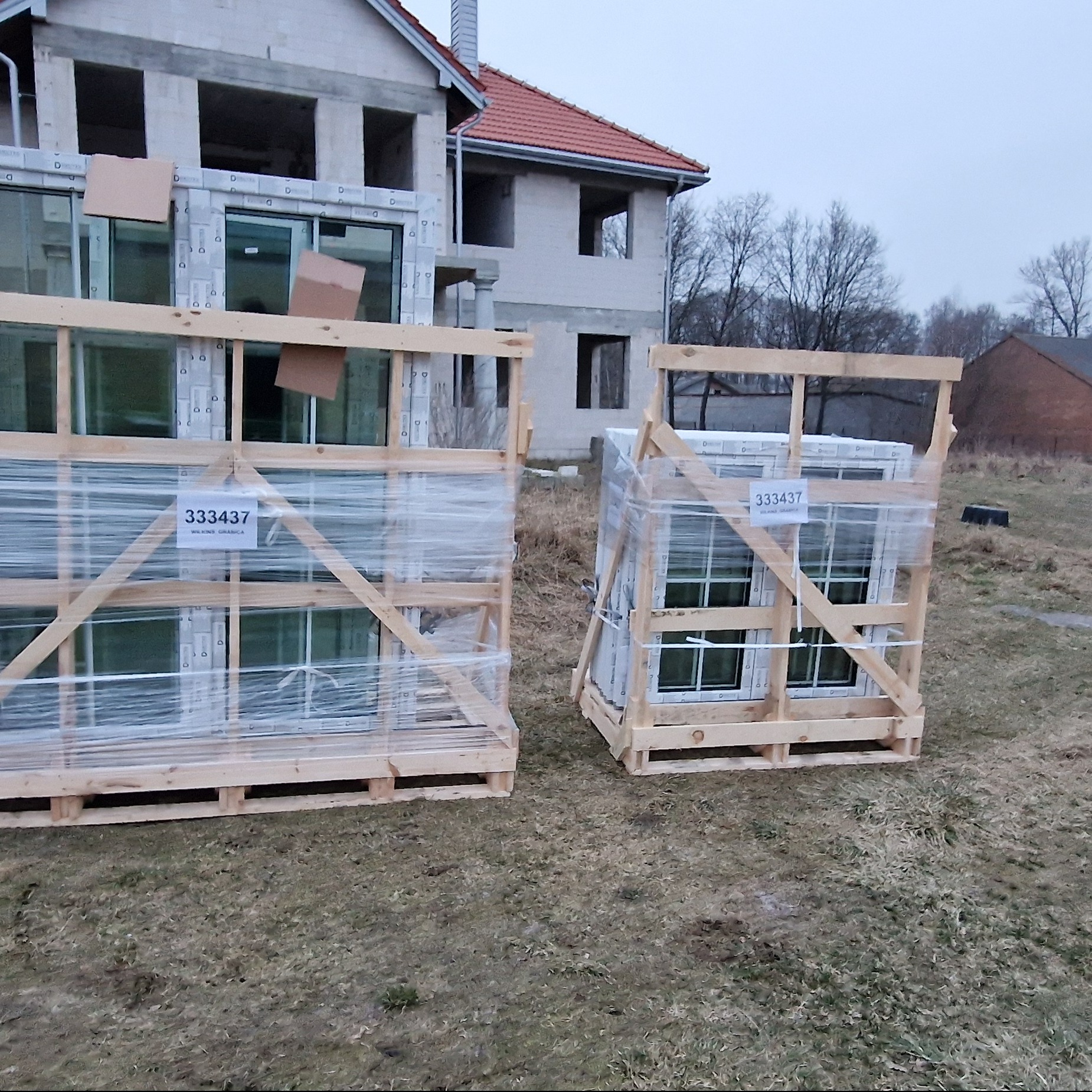 Dwa drewniane palety z zapakowanymi oknami PCV, zabezpieczone folią, stoją na trawie przed budynkiem w trakcie budowy.