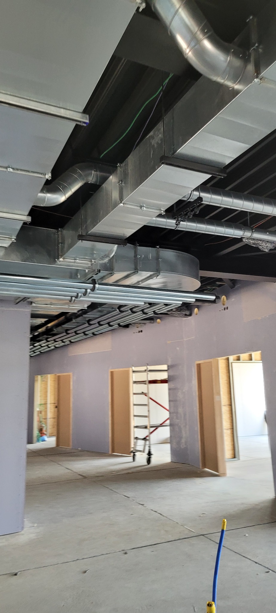 Wnętrze w trakcie wykańczania z widoczną instalacją wentylacyjną pod sufitem, rury wentylacyjne w kolorze srebrnym, ściany w kolorze jasnofioletowym, widoczne otwory drzwiowe i drabina na kółkach...