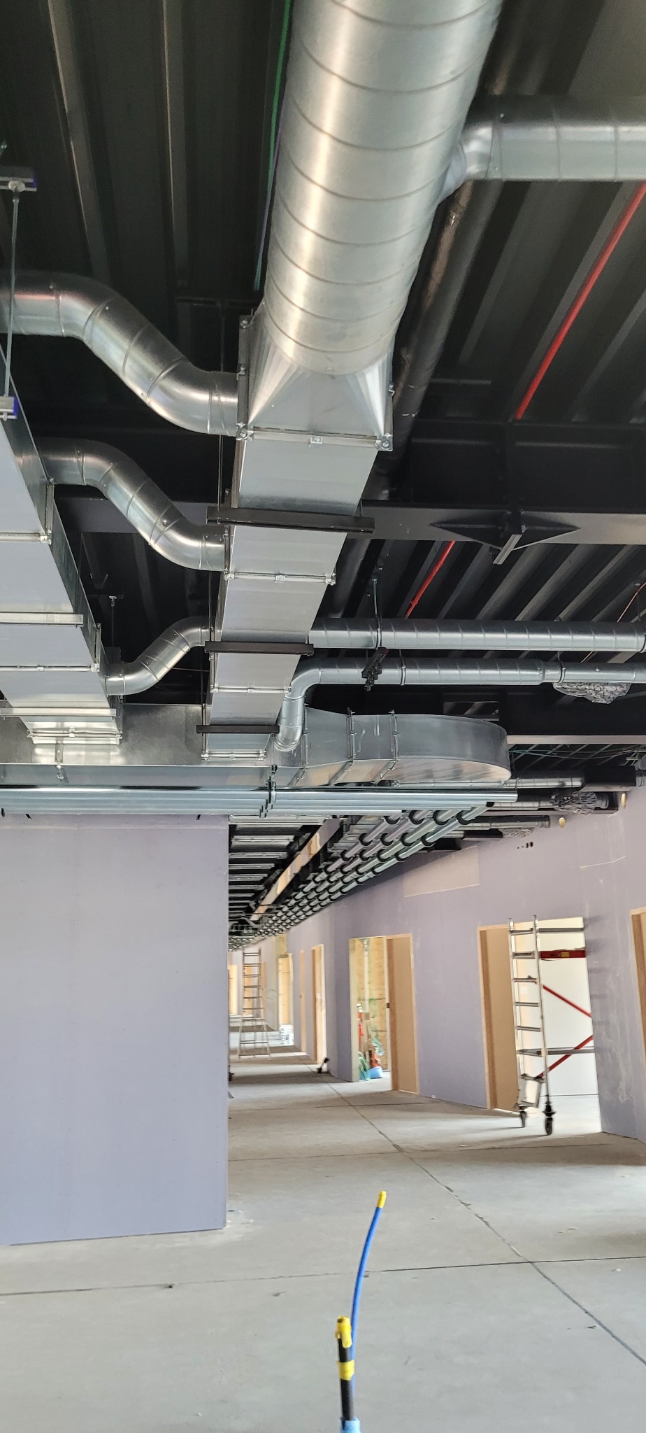 Widok na sufit z zainstalowanym systemem kanałów wentylacyjnych, rur i przewodów w surowym wnętrzu budynku, korytarz z widocznymi drabinami i otworami drzwiowymi w tle.