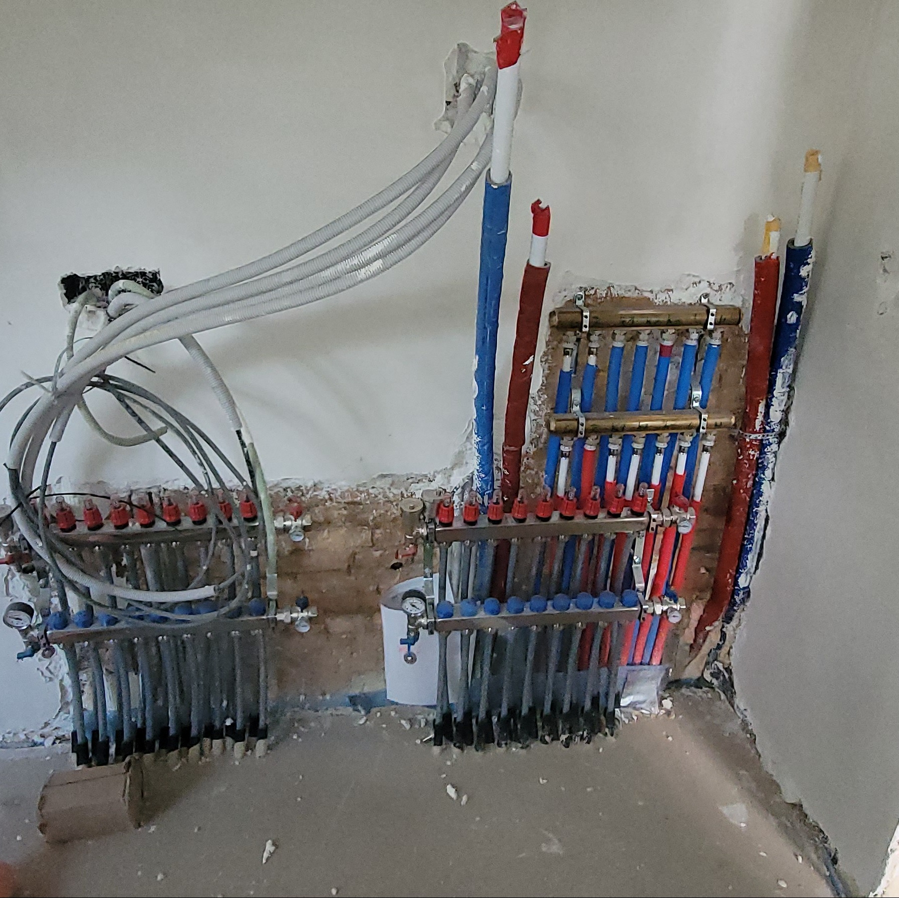 Rozdzielacz ogrzewania podłogowego z licznymi rurami w kolorach niebieskim i czerwonym, zamontowany w ścianie podczas remontu, widoczne przewody i manometry.