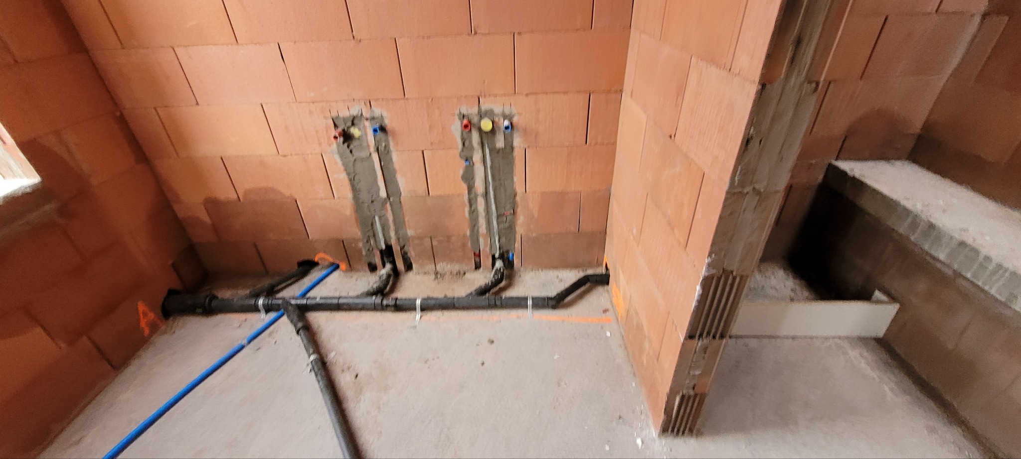 Surowe ściany z cegły z przygotowanymi otworami na instalację wodno-kanalizacyjną. Widoczne rury w kolorze czarnym i niebieskim, częściowo zamocowane do ściany. Na ścianie widoczne ślady po...