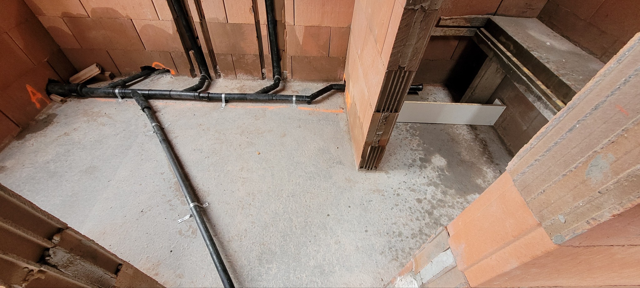 Surowy stan instalacji w budynku w trakcie budowy: widoczne czarne rury kanalizacyjne zamocowane do ściany z cegły dziurawki, z pomarańczowymi oznaczeniami na ścianach i podłodze.