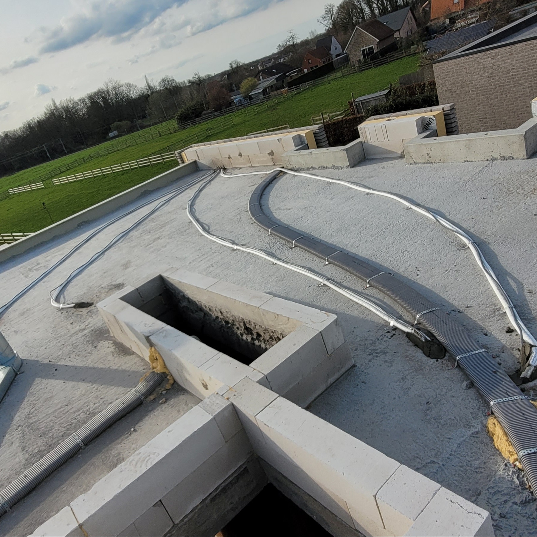 Instalacja przewodów wentylacyjnych na płaskim dachu budynku w trakcie budowy, widoczne przewody elastyczne w izolacji oraz fragmenty konstrukcji z bloczków betonowych i otwór wentylacyjny.