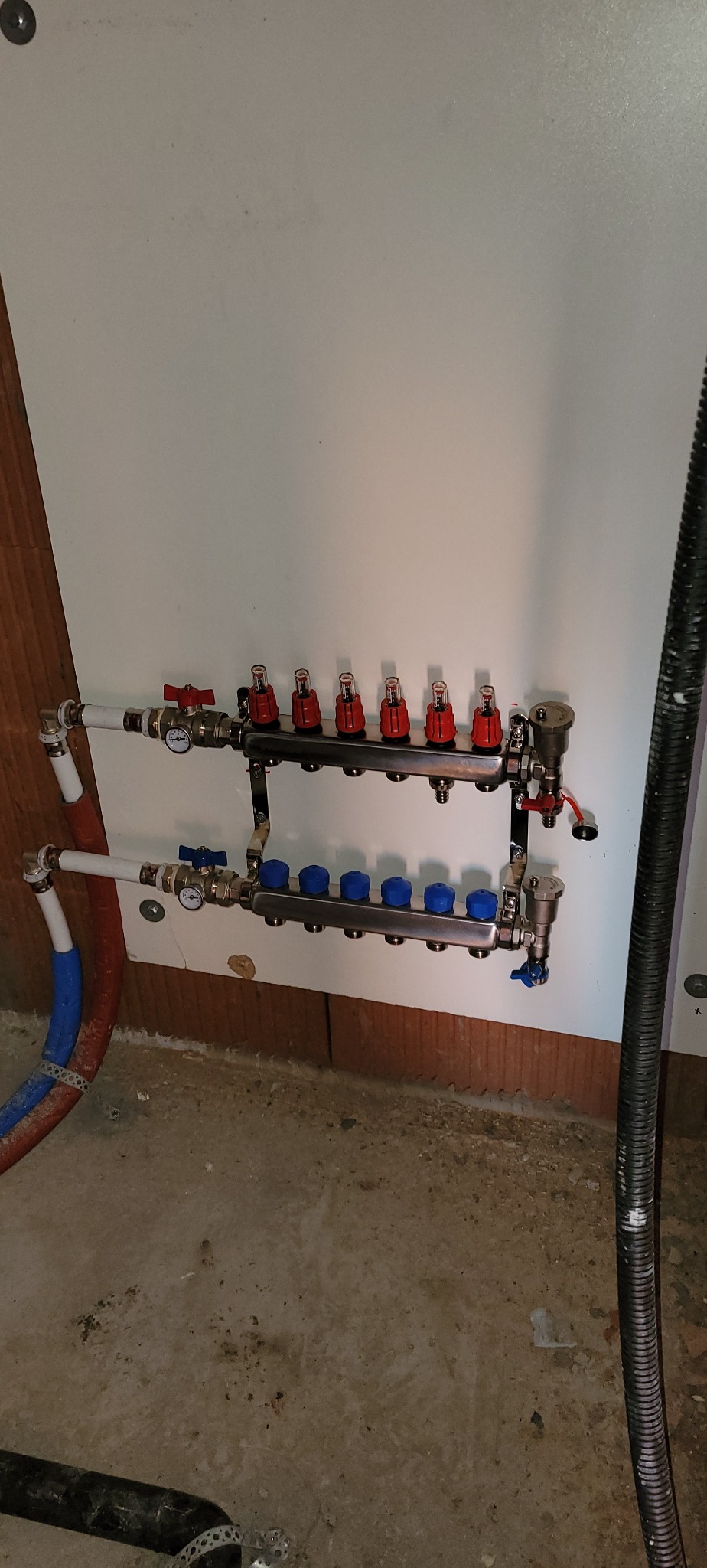 Rozdzielacz do ogrzewania podłogowego z czerwonymi i niebieskimi zaworami, zamontowany na ścianie w surowym wnętrzu.