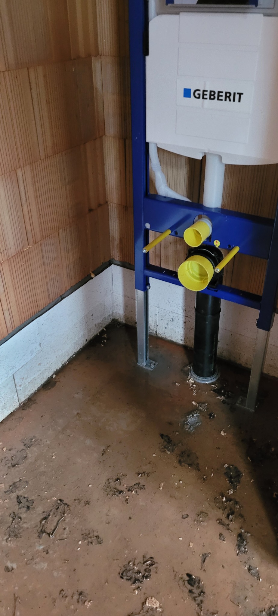 Stelaż podtynkowy z żółtymi elementami mocowany do ściany z cegły, widoczny odpływ kanalizacyjny i fragment brudnej posadzki.