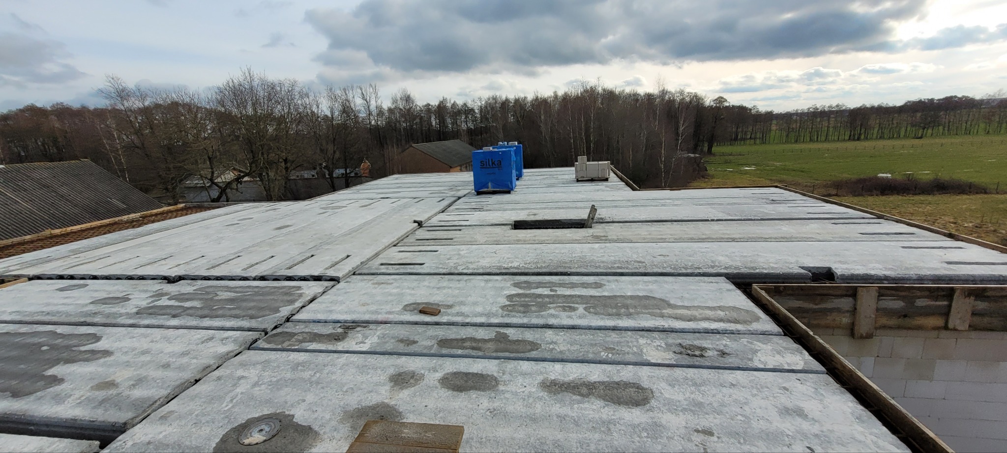 Widok z góry na strop Teriva z prefabrykowanych belek i pustaków betonowych, częściowo mokry, z widocznymi niebieskimi opakowaniami materiałów budowlanych i krajobrazem w tle.