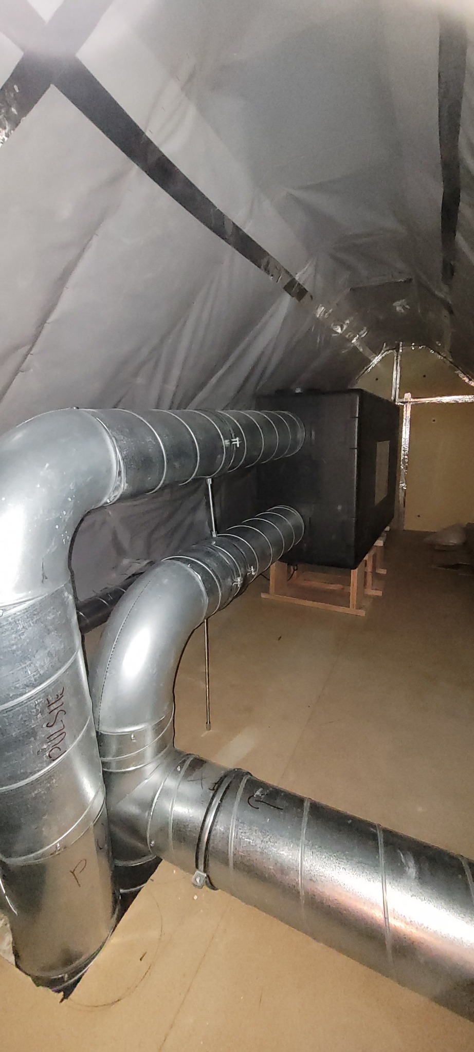 Czarny centralny element systemu wentylacyjnego z podłączonymi metalowymi rurami wentylacyjnymi w przestrzeni poddasza z izolacją.