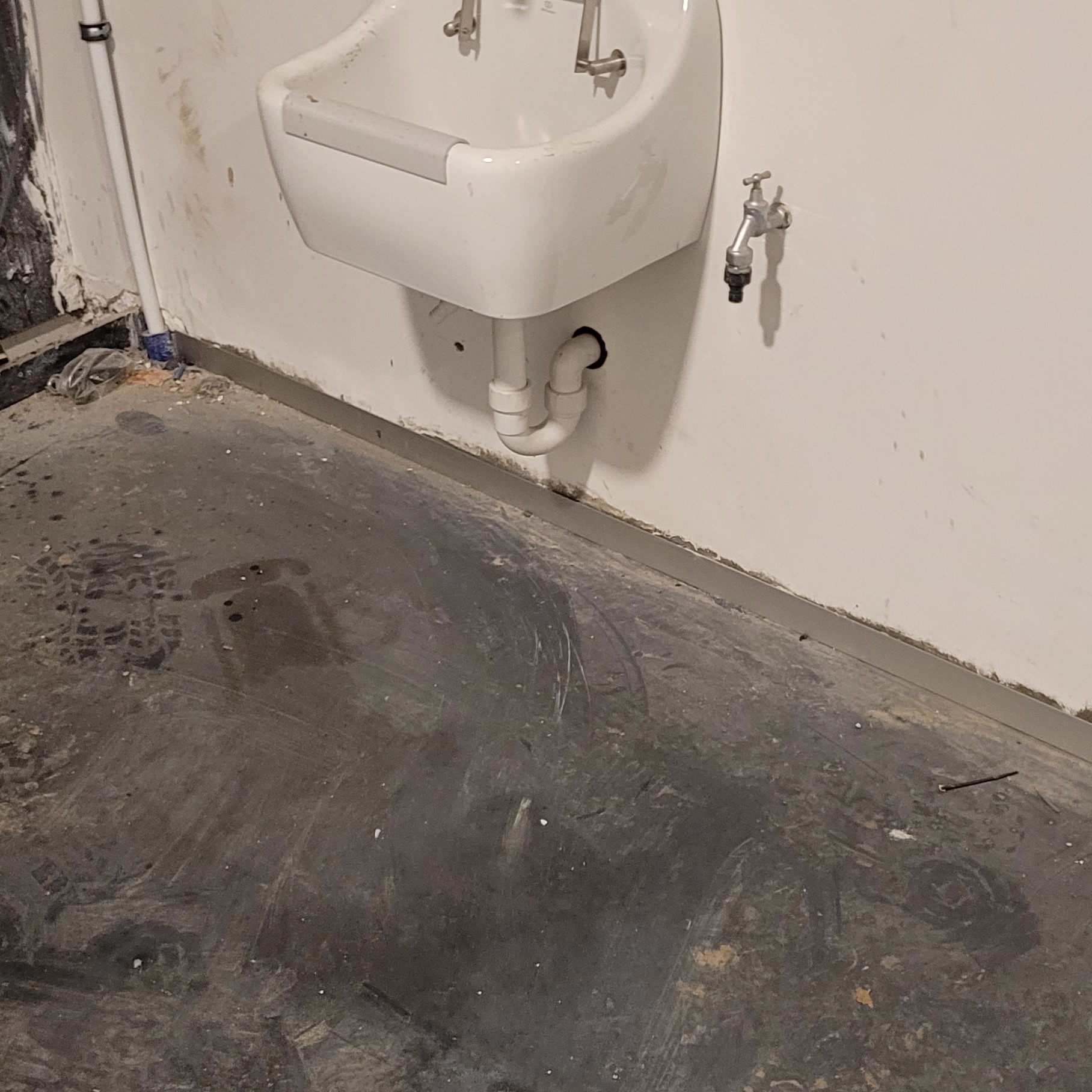 Montaż umywalki ceramicznej z widocznym syfonem i baterią ścienną w pomieszczeniu w trakcie wykończenia, ślady na podłodze.