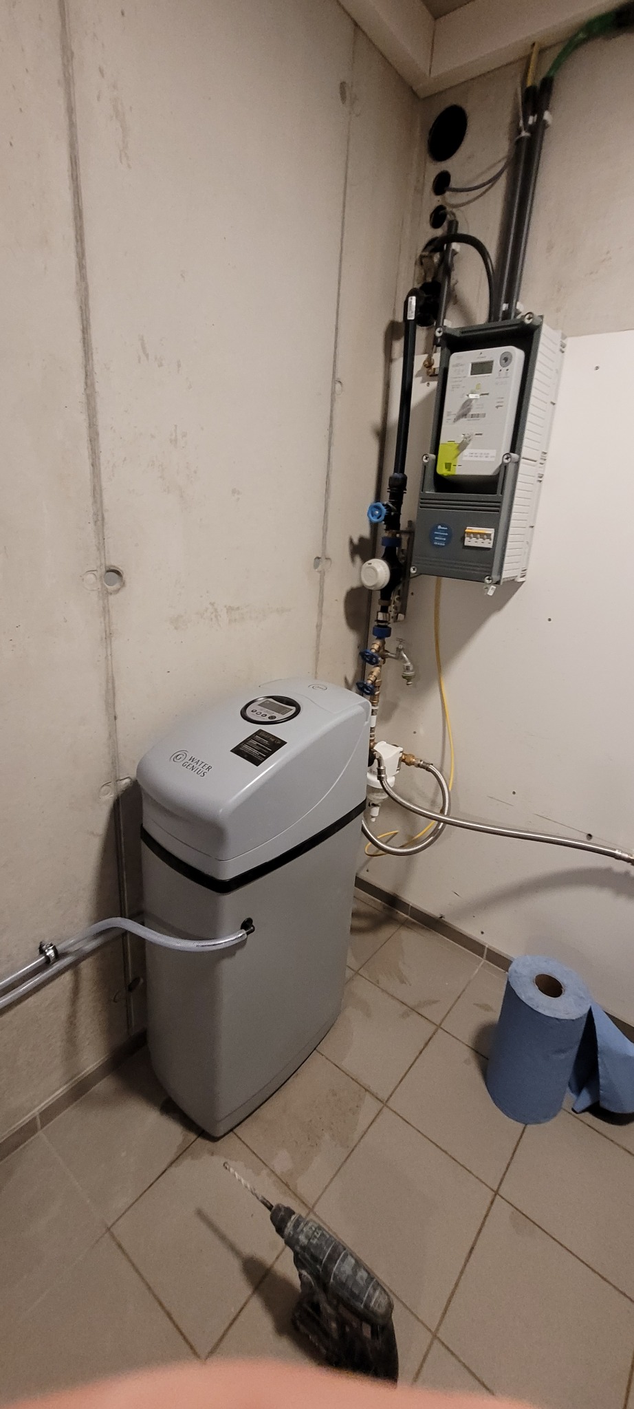 Szara stacja uzdatniania wody z podłączonymi rurami w pomieszczeniu gospodarczym, obok wiertarka akumulatorowa i rolka niebieskiego papieru toaletowego.