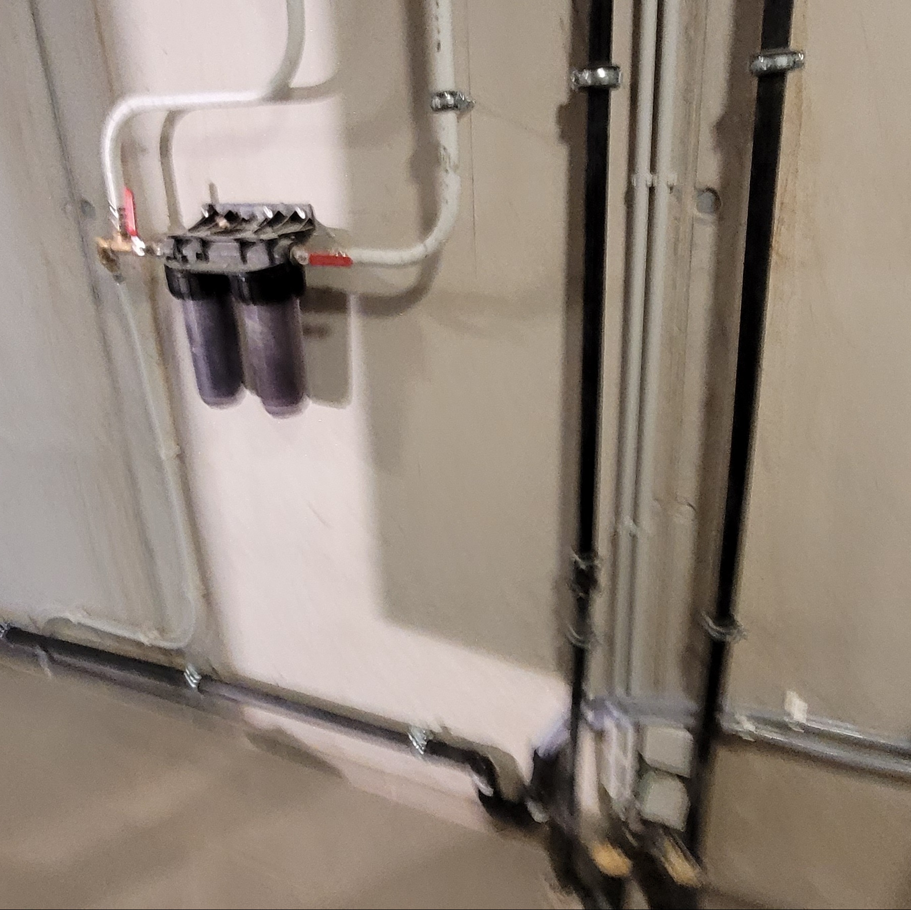 Zamontowany filtr wody z trzema przezroczystymi wkładami na ścianie, białe rury doprowadzające wodę, pionowe rury kanalizacyjne w tle.