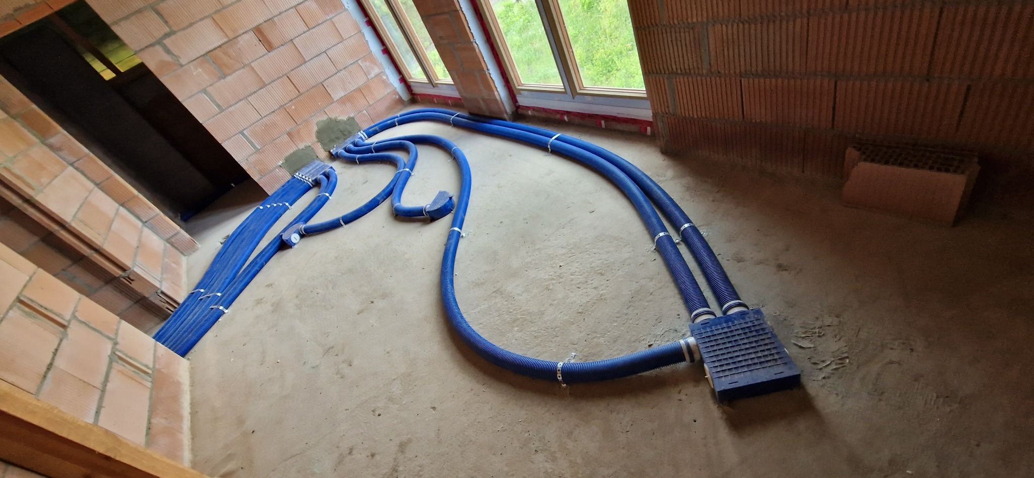 Niebieskie rury wentylacyjne w surowym wnętrzu z ceglanymi ścianami i oknami. Montaż instalacji w trakcie budowy. Widok z góry.