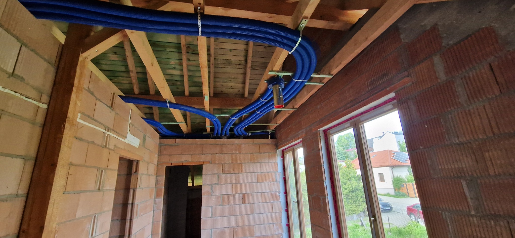 Niebieskie rury wentylacyjne przymocowane do drewnianej konstrukcji sufitu w surowym budynku z cegły. Widoczne okna i elementy instalacji.