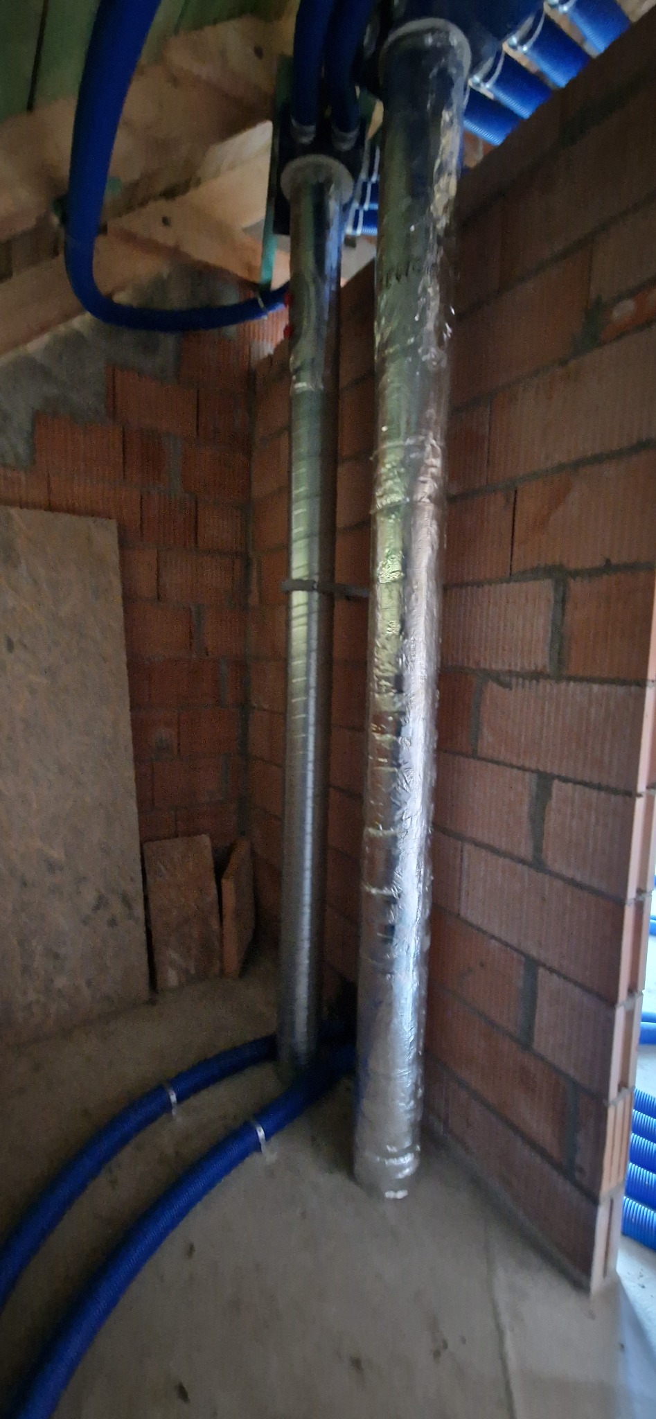 Dwie pionowe rury w srebrnej izolacji, część instalacji wentylacyjnej w surowym wnętrzu z ceglanymi ścianami i niebieskimi przewodami.