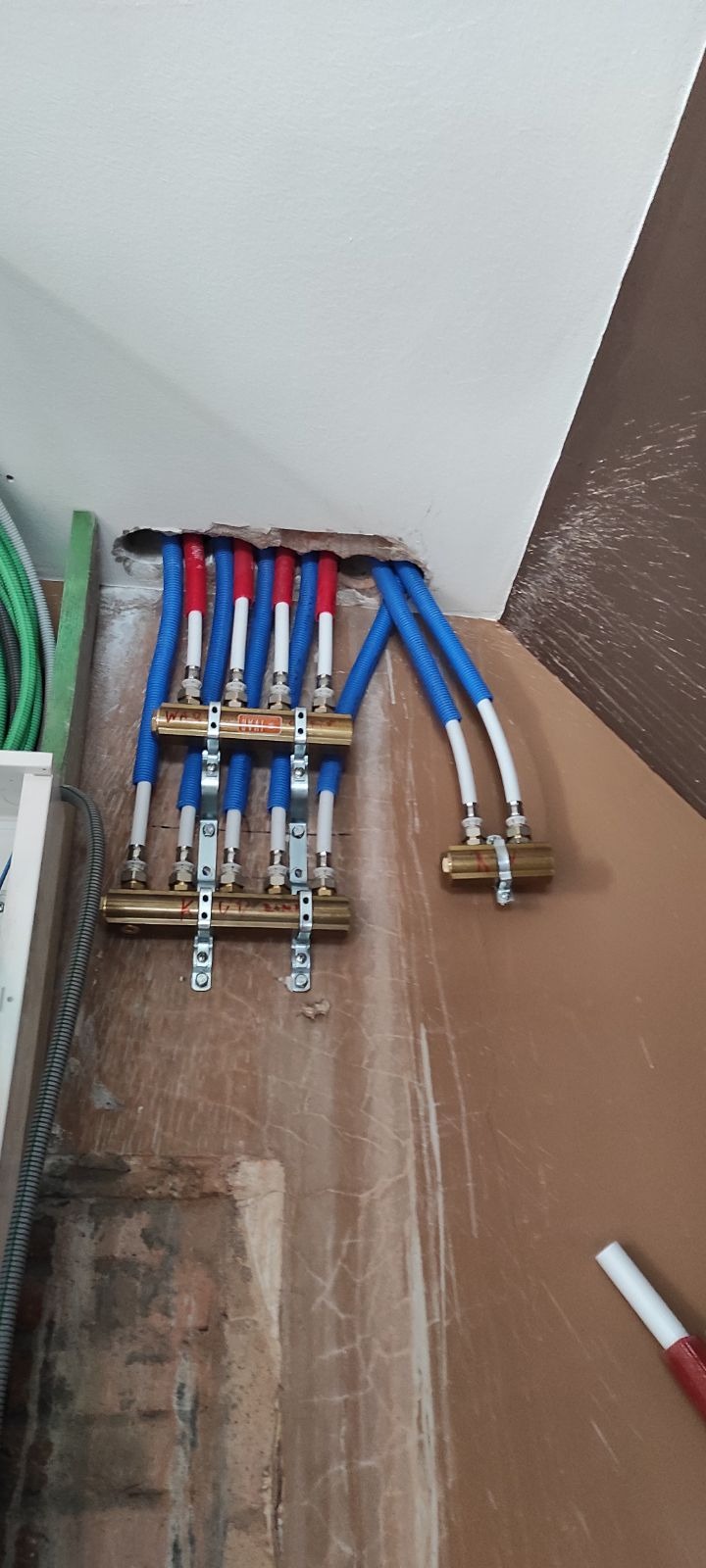 Rozdzielacz ogrzewania podłogowego z rurami PEX w kolorach: niebieskim, białym i czerwonym, zamontowany na ścianie w trakcie instalacji. Widoczne elementy mocujące.