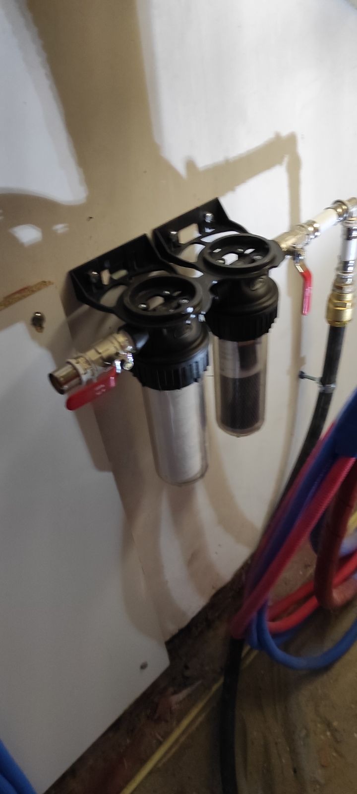 Filtr dwuczęściowy z zaworami, zamontowany na ścianie. Widoczne rury i przewody. Instalacja w pomieszczeniu gospodarczym.