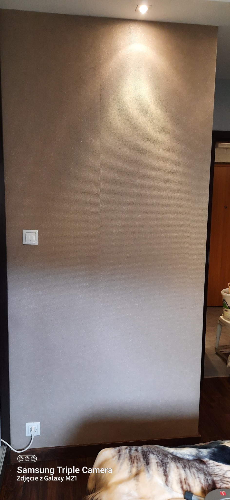 Ściana pokryta tapetą z delikatną teksturą, oświetlona punktowym światłem z góry, widoczny włącznik i gniazdko elektryczne.
