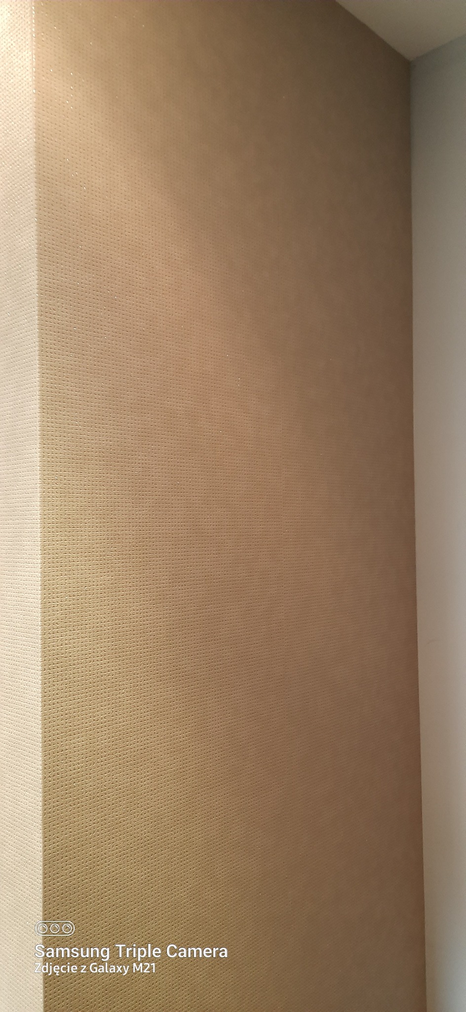 Beżowa tapeta o delikatnej strukturze na ścianie w rogu pokoju, sfotografowana z bliska z widocznym efektem ziarna.