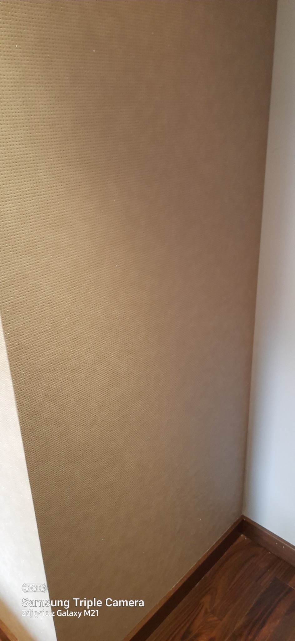 Fragment ściany pokrytej beżową tapetą o drobnej strukturze, widoczny kąt pomieszczenia z drewnianą listwą przypodłogową.