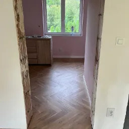 Układanie parkietu w jodełkę w pokoju z różowymi ścianami, widoczne surowe ościeżnice i okno z widokiem na zieleń.