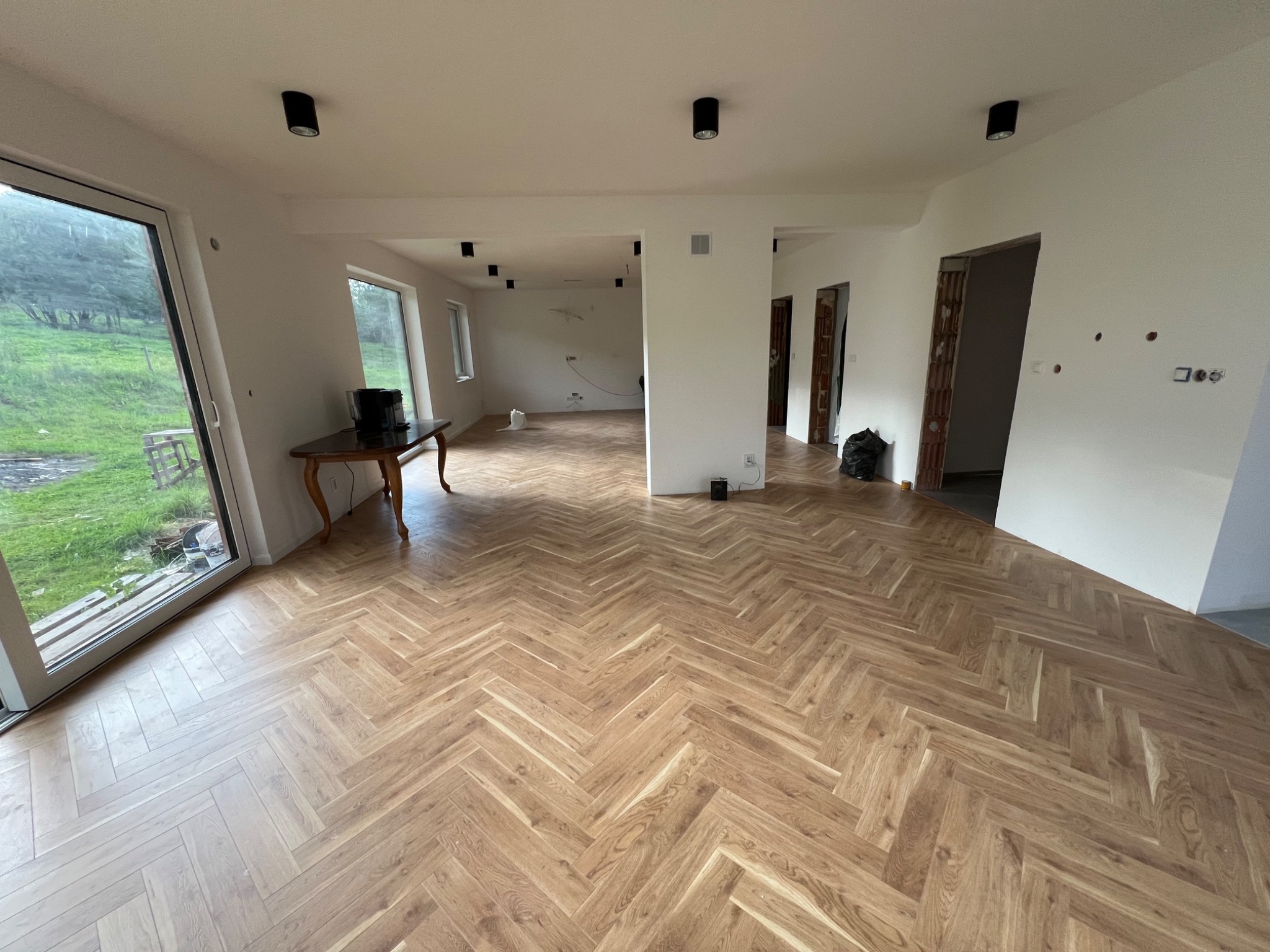 Jasne panele ułożone w jodełkę w przestronnym, częściowo wykończonym wnętrzu z białymi ścianami, widoczne punkty świetlne na suficie, stolik przy oknie i otwarte przejścia między pomieszczeniami.