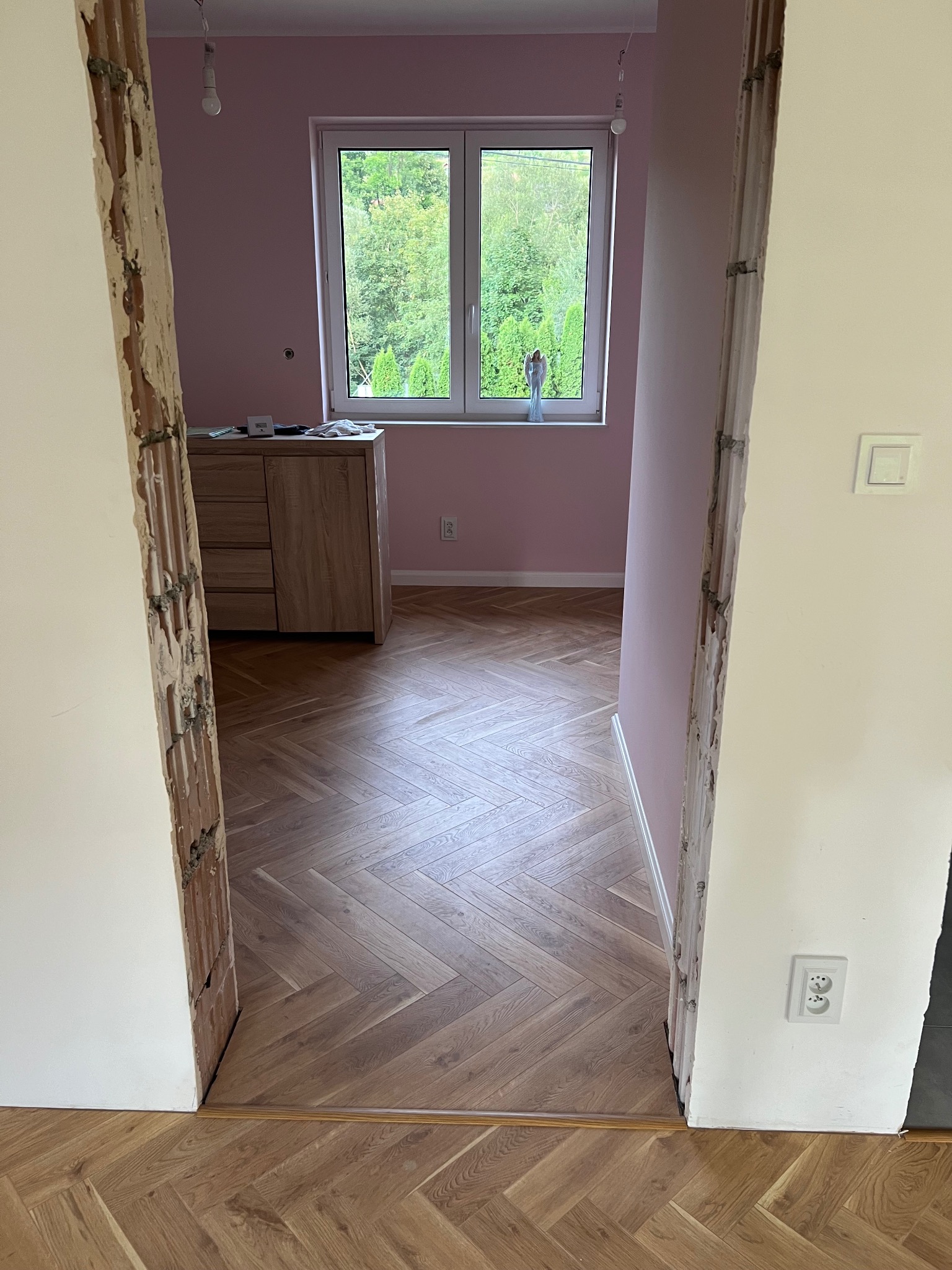 Układanie parkietu w jodełkę w pokoju z różowymi ścianami, widoczne surowe ościeżnice i okno z widokiem na zieleń.