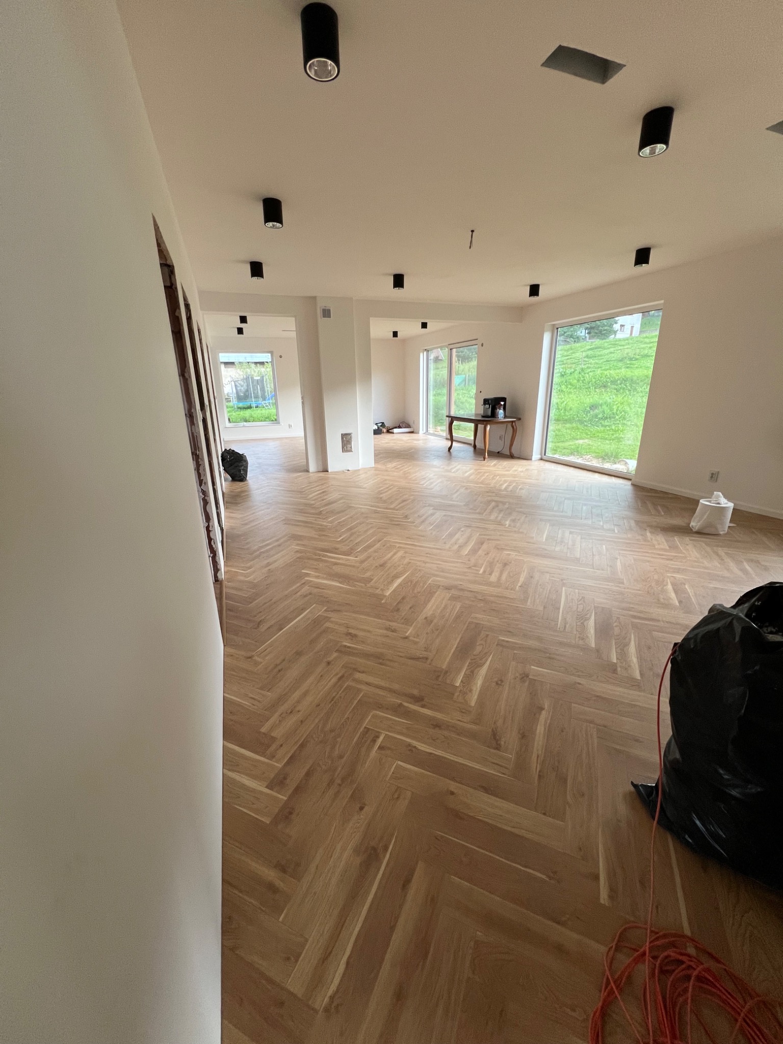 Układanie parkietu w jodełkę w przestronnym, jasnym wnętrzu. Widoczne czarne worki na śmieci i zwój pomarańczowego kabla, co sugeruje trwające prace wykończeniowe. Okna z widokiem na zieleń...