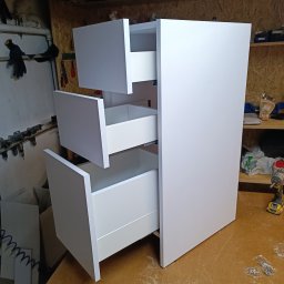 Stolkra - Biała szafka z trzema wysuniętymi szufladami, widoczne narzędzia montażowe i wkrętarka na drewnianym blacie w warsztacie. Detal wykończenia mebla.