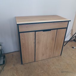 Stolkra - Drewniana szafka z trzema drzwiczkami i szufladą, czarne obramowanie, widoczna struktura drewna, na podłodze z płyt OSB, tło białe.