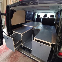 Stolkra - Zabudowa kampera z szarej sklejki: wysuwane szafki, blat stołu na metalowej nodze. Widok z otwartych drzwi bagażnika, wnętrze pojazdu.