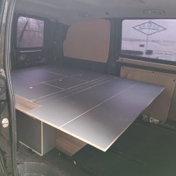 Stolkra - Wnętrze busa z zamontowaną zabudową meblową, szara płyta meblowa na drewnianej konstrukcji. Widok z otwartych tylnych drzwi pojazdu.