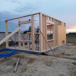 Stolkra - Wznoszenie drewnianej konstrukcji szkieletowej domu na fundamencie, widoczne elementy rusztowania i niebieski przewód. Budowa w toku, tło: zachodzące słońce.