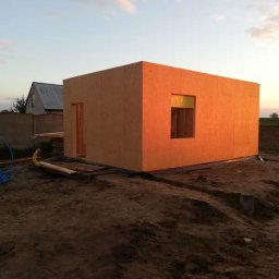 Stolkra - Wczesny etap budowy domu szkieletowego z widoczną konstrukcją ścian z płyty OSB, otworem okiennym i drzwiowym na tle zachodzącego słońca.