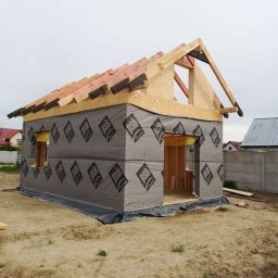 Stolkra - Surowy stan domu szkieletowego z widoczną konstrukcją dachu i membraną na ścianach, na tle działki z gruntem. W tle zabudowa jednorodzinna.